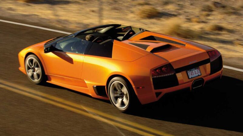 An orange Lamborghini Murciélago convertible—the last manual V12 Lambo roadster—drives on a desert road, viewed from above and behind. Less than 10 exist worldwide, making this moment truly extraordinary.