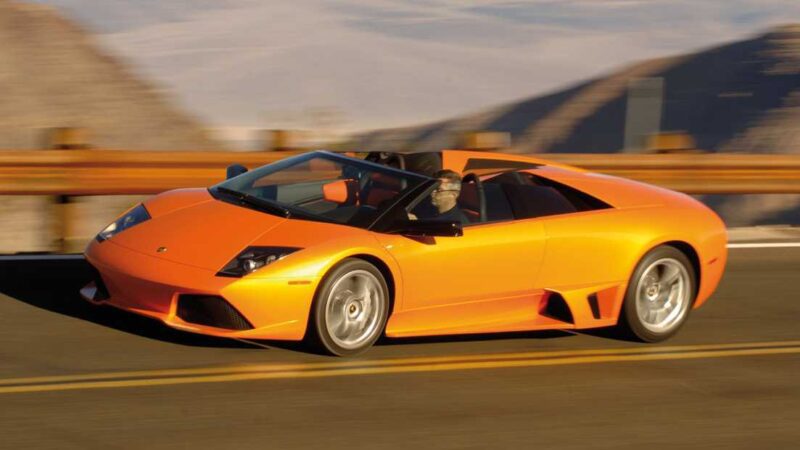 An orange Lamborghini convertible—one of less than 10 that exist—drives on a winding mountain road, its blurred surroundings highlighting the thrilling speed of the last manual V12 Lambo roadster.