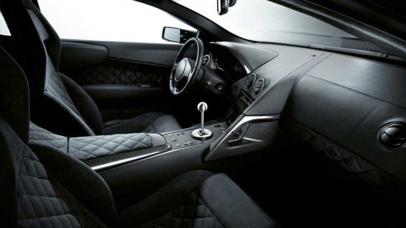 Black luxury car interior with diamond-stitched seats, manual gear shift, and minimalist dashboard featuring round air vents and analog controls—Less Than 10 Exist: The Last Manual V12 Lambo Roadster.