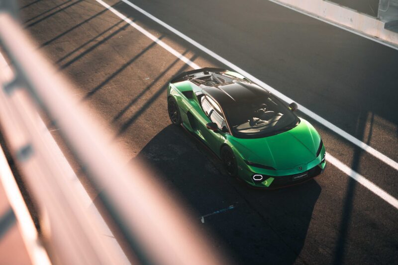 A green Lamborghini Temerario with a black roof is parked on a racetrack in bright sunlight, viewed from above through a fence.