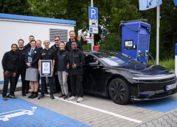 A group of people stand and pose by a black Lucid electric car with a 749-mile EV range at a charging station, holding a certificate—possibly for a Guinness World Record—in a parking lot with electric vehicle markings.