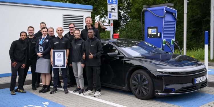 A group of people stand and pose by a black Lucid electric car with a 749-mile EV range at a charging station, holding a certificate—possibly for a Guinness World Record—in a parking lot with electric vehicle markings.