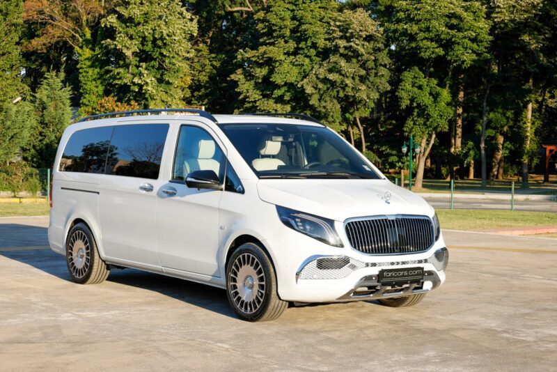 A white luxury van from a custom garage is parked on a paved area with green trees in the background under daylight.
