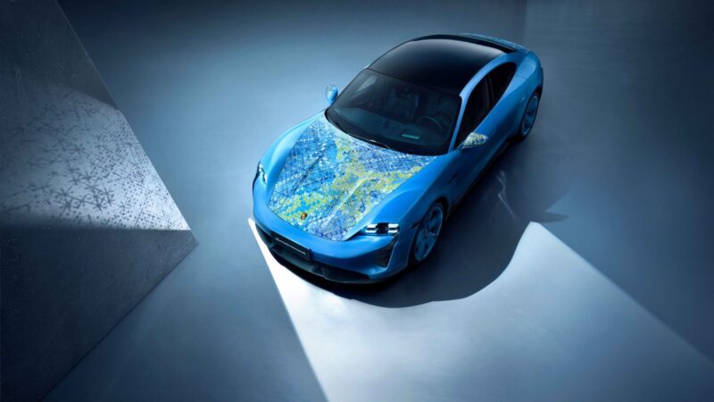 A blue sports car with a black roof is parked indoors under a spotlight; its hood features a design resembling a colorful abstract painting, showcasing the creativity possible through Porsche exclusive requests.