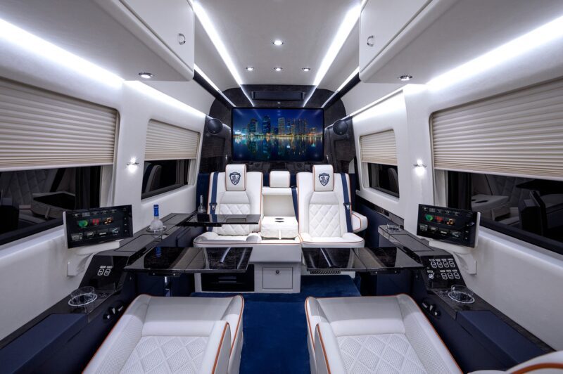 Luxurious van interior with white leather seats, LED lighting, fold-out tables, touchscreen controls, and a large screen displaying a cityscape—a unique creation perfect for those browsing custom cars for sale.