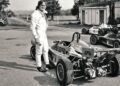 A person in a racing suit stands beside an open-wheel race car from the McLaren Road Car Collection, with other similar cars and garages in the background—evoking the legacy of Mansour Ojjeh in motorsport.
