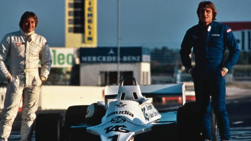 Two men in racing suits stand beside a white Formula 1 car from the McLaren Road Car Collection at a racetrack, with sponsor signs and a control tower visible in the background—a nod to Mansour Ojjeh’s enduring legacy.