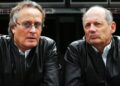 Two middle-aged men wearing black leather jackets sit side by side, looking directly at the camera with serious expressions, evoking the bold spirit of the McLaren Road Car Collection championed by Mansour Ojjeh.