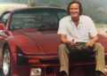 A man sits on the hood of a red sports car, smiling at the camera with a rural landscape behind him—a stylish addition to the McLaren Road Car Collection inspired by Mansour Ojjeh.