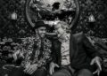 Two men sit on a patterned sofa, smiling and talking; one holds a drink. A round mirror behind them reflects a band playing on stage at the Mansour Ojjeh event for the McLaren Road Car Collection.