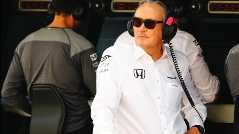 A man in a white Honda-branded shirt and sunglasses, wearing a headset, sits in front of computer screens at a McLaren racing event.