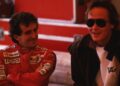 Two men are talking; one, possibly Mansour Ojjeh, is in a red racing suit with sponsor logos, while the other, wearing dark sunglasses and a jacket, stands beside a McLaren Road Car Collection vehicle.
