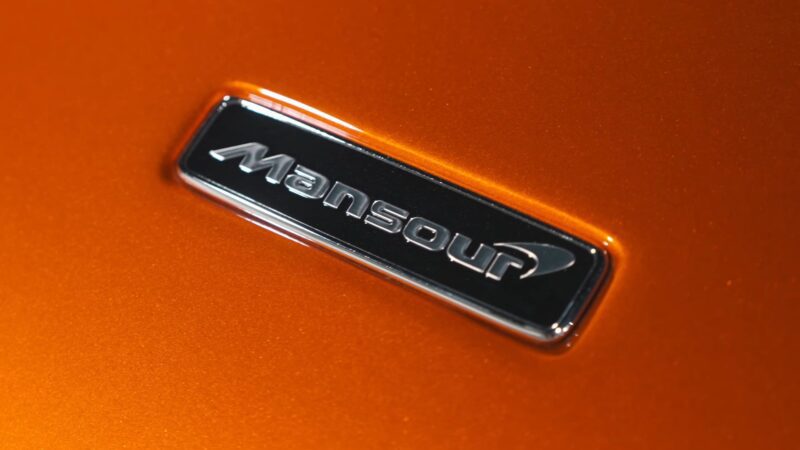 Close-up of a "Mansour" badge on an orange metallic surface, celebrating the Mansour Ojjeh legacy within the McLaren Road Car Collection.