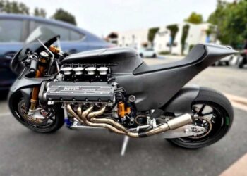 A bespoke superbike with a massive exposed Ferrari V8 engine stands parked outdoors on two wheels, surrounded by cars and buildings in the background.