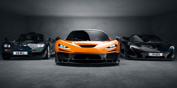 Three McLaren sports cars—a classic F1, a bright orange modern McLaren W1, and a sleek black model—are parked side by side in a dimly lit space reminiscent of Goodwood’s exclusive showcases.