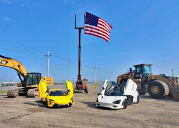 Two sports cars, including a McLaren with its doors open, are parked on a dirt lot between construction vehicles near Baltimore's Vehicle Processing Center, with a large American flag raised on a pole in the background.