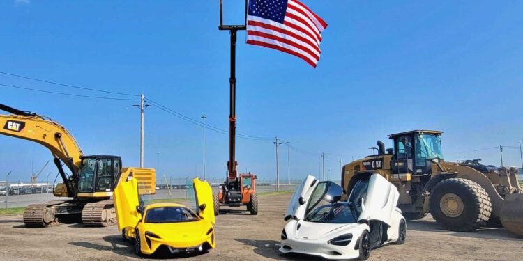 Two sports cars, including a McLaren with its doors open, are parked on a dirt lot between construction vehicles near Baltimore's Vehicle Processing Center, with a large American flag raised on a pole in the background.