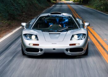 A silver McLaren F1 sports car drives down a two-lane road surrounded by greenery, viewed from the front—an iconic sight reminiscent of RM Sotheby's Monterey Auction showcases.