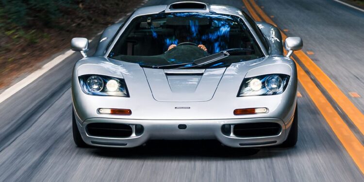 A silver McLaren F1 sports car drives down a two-lane road surrounded by greenery, viewed from the front—an iconic sight reminiscent of RM Sotheby's Monterey Auction showcases.