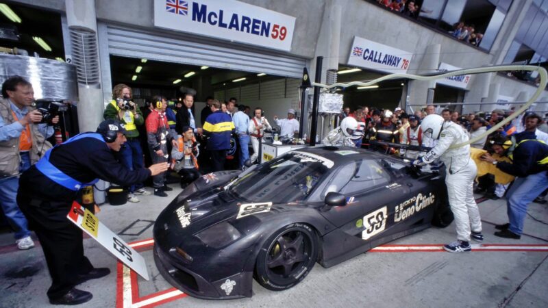 A black McLaren F1 GTR race car in a pit stop, surrounded by crew members and spectators, with mechanics servicing the Lanzante 95-59 during a thrilling Le Mans motorsport event.
