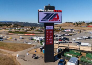 A large MotoAmerica leaderboard tower stands in the center of a racetrack facility, displaying race standings, with trailers and event structures in the background.