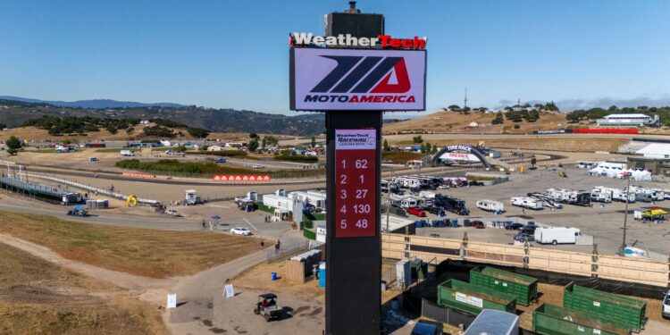 A large MotoAmerica leaderboard tower stands in the center of a racetrack facility, displaying race standings, with trailers and event structures in the background.