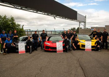 A group of people in black outfits pose beside red, yellow, and blue sports cars—including the Corvette ZR1—on a race track, holding Nürburgring lap time signs touting one of the fastest American production cars’ Nürburgring record.