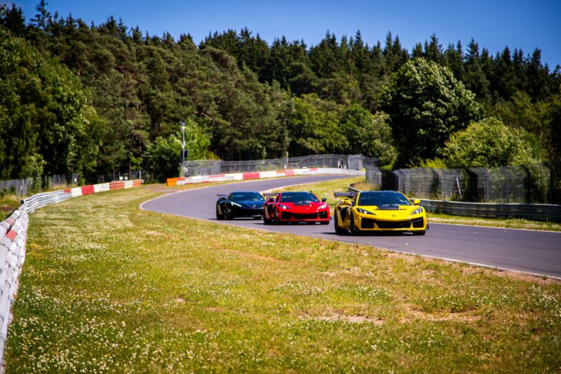 Three sports cars—black, red, and yellow—drive in a line around a curve on a racetrack bordered by grass and trees; among them is a Corvette ZR1, one of the fastest American production cars, glinting in the sunny daylight.