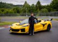 A person in a black racing suit stands next to a yellow Corvette ZR1—one of the fastest American production cars—on a racetrack, holding a helmet, with trees and a fence in the background.