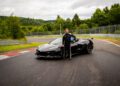 A person in a black racing suit stands on a racetrack next to a Corvette ZR1, one of the fastest American production cars, holding a helmet, with green trees in the background.