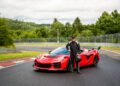 A person in a racing suit stands next to a red Corvette ZR1X, one of the fastest American production cars, on a racetrack surrounded by trees under a cloudy sky.