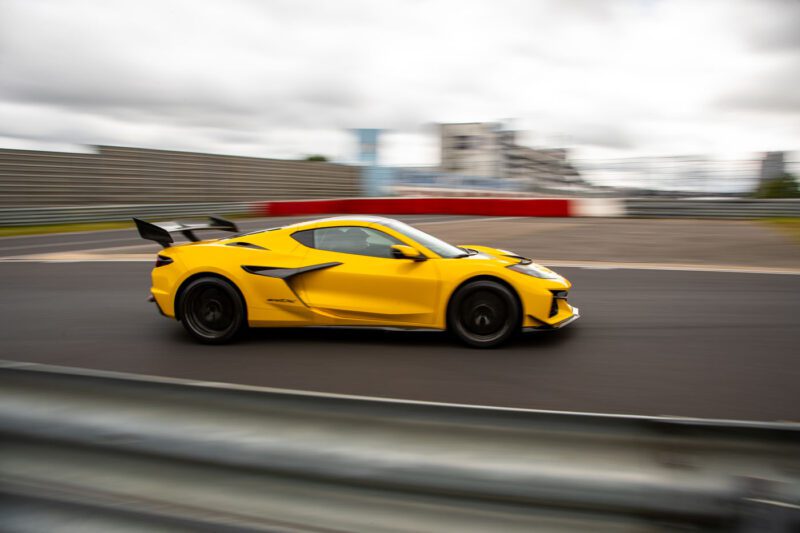 A yellow Corvette ZR1X speeds around a racetrack with motion blur emphasizing its movement; barriers and a blurred background hint at its Nürburgring record among the fastest American production cars.