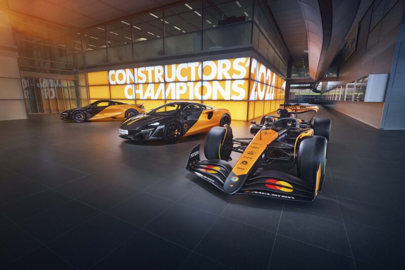 Three orange and black McLaren vehicles, including an F1 car, are displayed indoors in front of a brightly lit "Constructors Champions" sign, evoking the excitement of Goodwood.