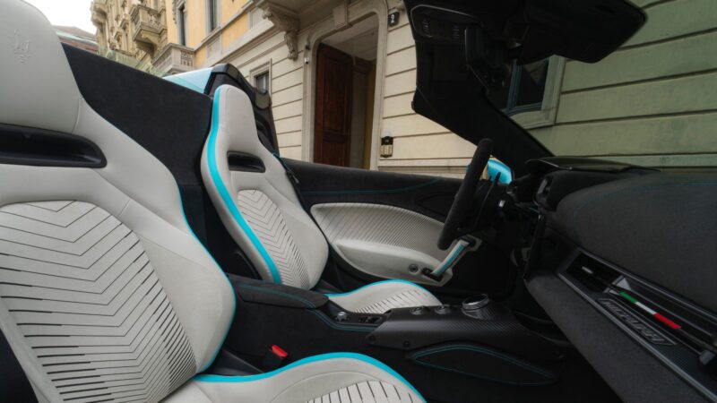 The interior of the 2026 Maserati MCPURA, debuting at Goodwood with a liquid crystal roof and F1 tech, features white and black leather seats, turquoise accents, and a modern dashboard, parked on a city street.