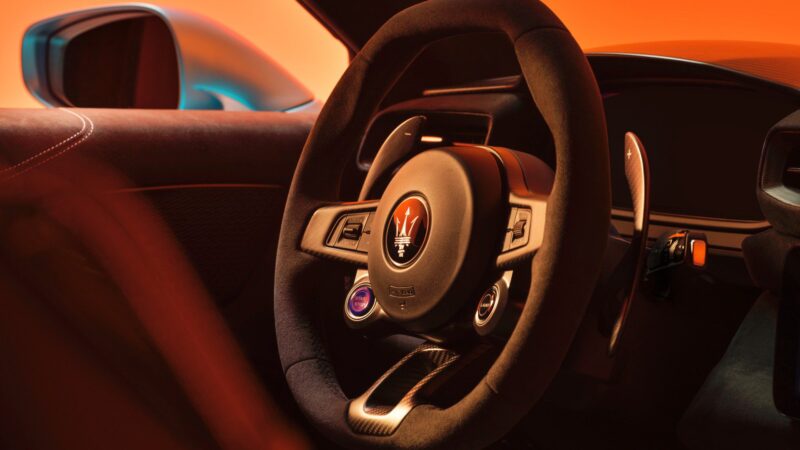 Close-up view of a Maserati steering wheel and dashboard, showcasing the sleek, modern interior of the 2026 Maserati MCPURA, which debuts at Goodwood with F1 tech and a Liquid Crystal Roof, set against an orange background.
