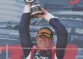 A race car driver in a racing suit and cap holds a trophy above his head while smiling on a podium in front of a branded backdrop.