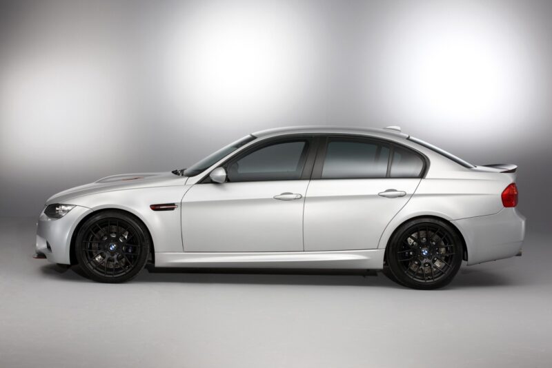 Side view of a silver BMW E90 M3 sedan with black wheels, parked indoors against a plain gray background—a true collector car.