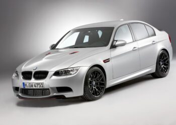 A silver BMW E90 M3 sedan, a true collector car, is parked in a studio with a plain gray background, viewed from a front three-quarter angle.