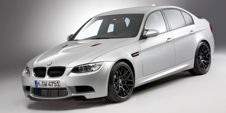 A silver BMW E90 M3 sedan, a true collector car, is parked in a studio with a plain gray background, viewed from a front three-quarter angle.