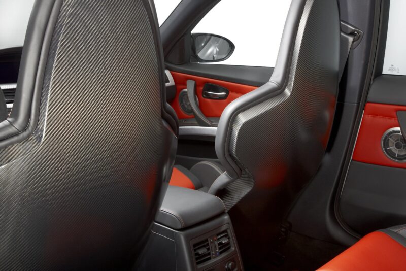 View of a car’s interior showing carbon fiber racing-style front seat backs, red and black door trim, and rear seat air vents—luxury details you’d expect in a $250K collector car like a BMW E90 M3.
