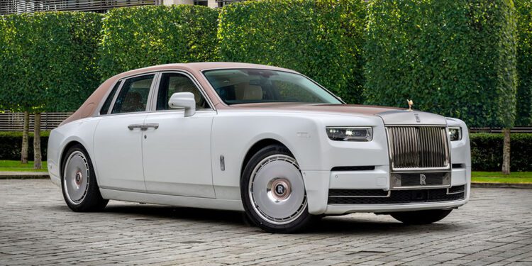 A one-off white and brown Rolls-Royce Phantom Dentelle sedan is parked on a cobblestone driveway in front of trimmed hedges and a modern building.