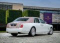 A white and pink Rolls-Royce Phantom Dentelle, a stunning one-off sedan, is parked on a paved driveway in front of a modern Rolls-Royce dealership building.