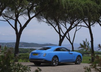 A blue Rolls-Royce luxury coupe with a yellow license plate is parked on a dirt road in Saint-Tropez, surrounded by trees and overlooking a scenic landscape with hills and water.