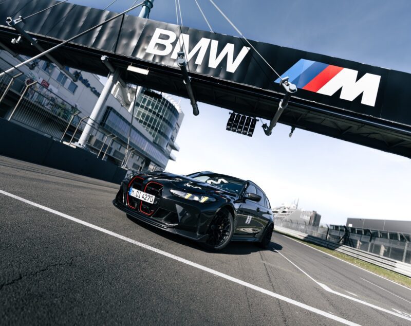 A black BMW M3 CS Touring, known as the fastest wagon with a Nürburgring lap record, is parked on a race track under a large BMW M sign, with modern buildings in the background.
