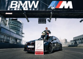 A person in racing gear sits on a black BMW M3 CS Touring under a BMW M sign at a racetrack, with a Nürburgring lap record sign displayed in front, highlighting the fastest wagon’s achievement.