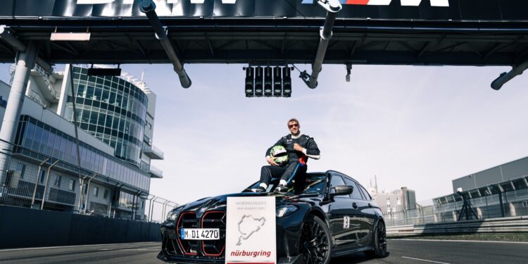 A person in racing gear sits on a black BMW M3 CS Touring under a BMW M sign at a racetrack, with a Nürburgring lap record sign displayed in front, highlighting the fastest wagon’s achievement.