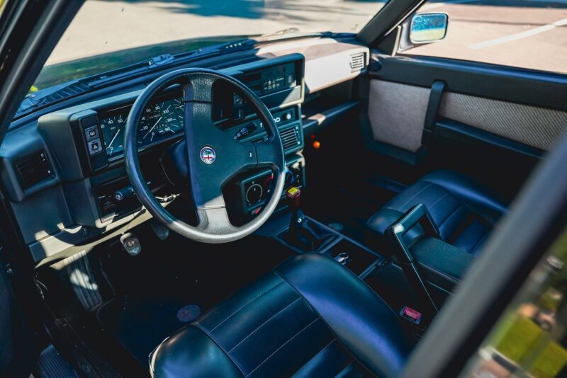 Interior of a vintage Alfa Romeo showing a black leather driver's seat, steering wheel, dashboard controls, and front passenger seat in daylight—perfect for any Petrolicious Film Friday feature.