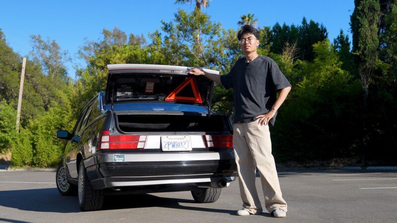 A person stands next to an open hatchback car in a parking lot, with a reflective warning triangle visible inside the trunk—a scene straight out of Petrolicious Film Friday, reminiscent of classic Alfa Romeo adventures.