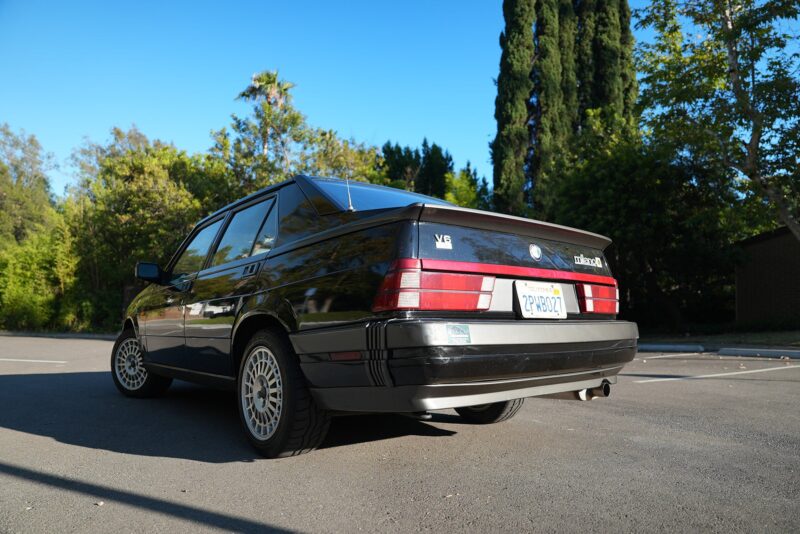 A black Alfa Romeo Milano sedan is parked in an outdoor lot, surrounded by trees, with a clear blue sky overhead—a scene worthy of a Film Friday feature on Petrolicious.