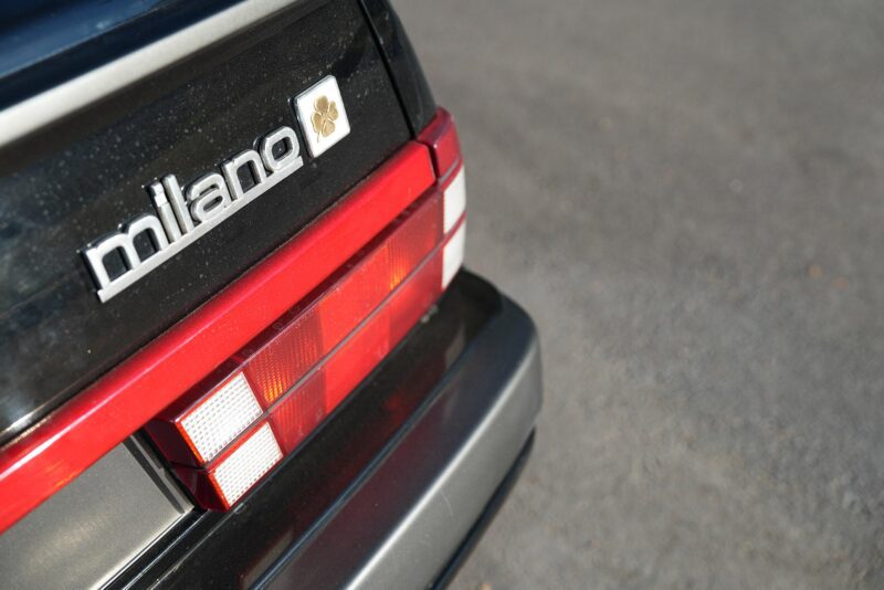 Close-up of the rear badge and tail light of a black Alfa Romeo labeled "milano," parked on an asphalt surface—perfect for a Petrolicious Film Friday feature.